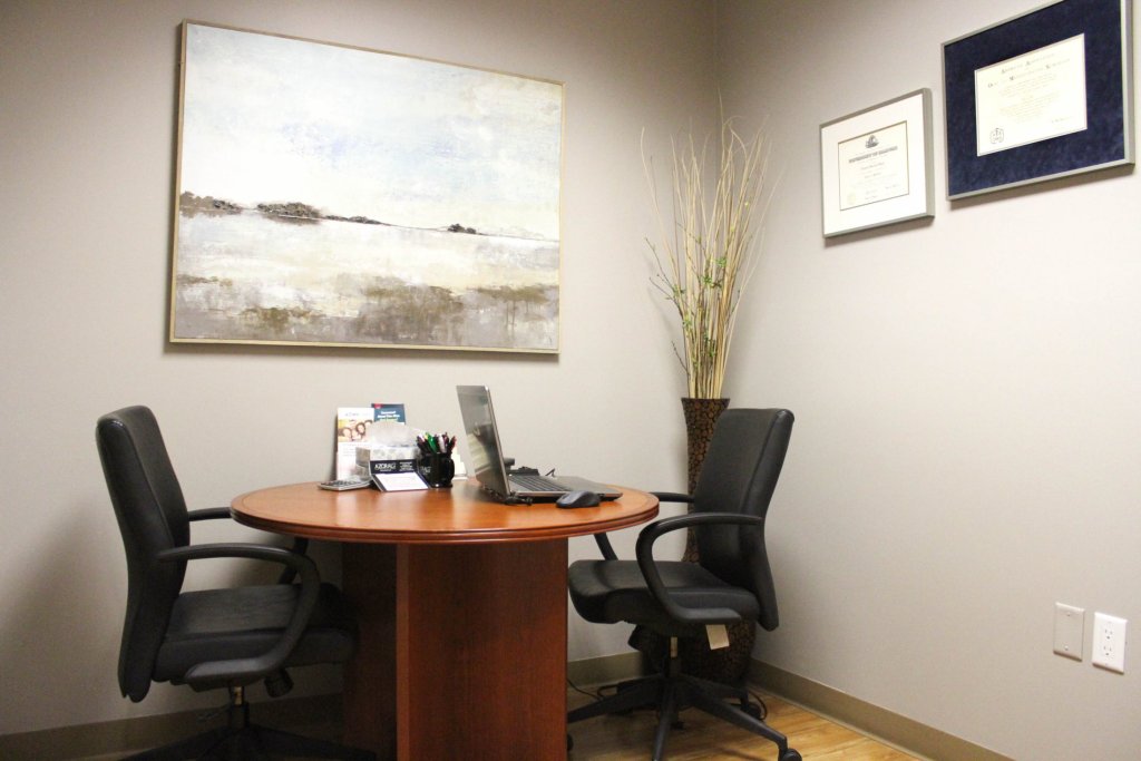 Small office with round table, two chairs, laptop, painting, and framed certificates