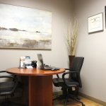 Small office with round table, two chairs, laptop, painting, and framed certificates