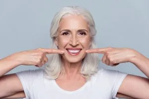 Older woman with gray hair pointing to her teeth with both hands